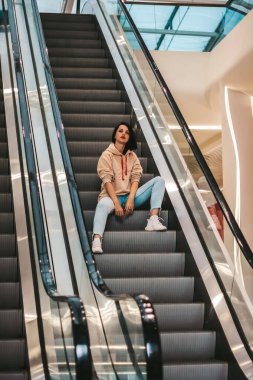 Kız yürüyen merdivende otururken ata biniyor. Alışveriş merkezi, güzel esmer spor tarzında..