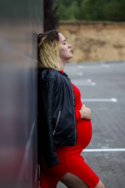 Jovem grávida em um vestido vermelho no estacionamento. gravidez é uma alegria. barriga grande grávida. nova vida. — Fotografia de Stock