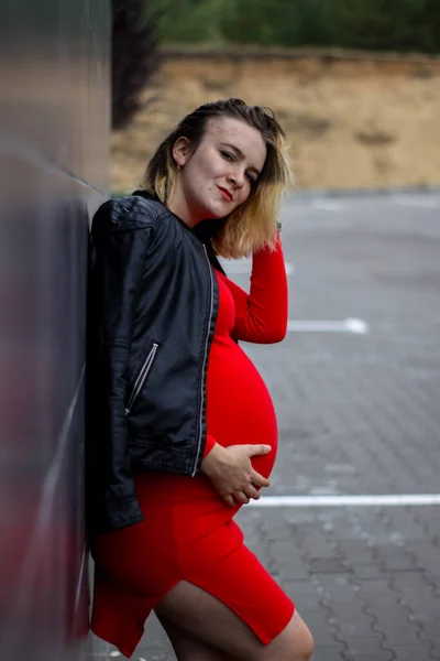 Giovane ragazza incinta in un vestito rosso nel parcheggio. la gravidanza è una gioia. pancia grande incinta. nuova vita. — Foto Stock