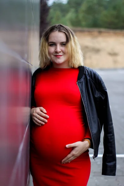 Giovane ragazza incinta in un vestito rosso nel parcheggio. la gravidanza è una gioia. pancia grande incinta. nuova vita. — Foto Stock