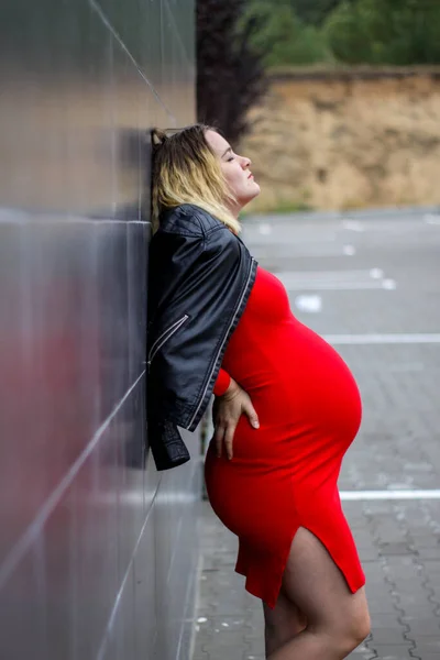 Jovem grávida em um vestido vermelho no estacionamento. gravidez é uma alegria. barriga grande grávida. nova vida. — Fotografia de Stock