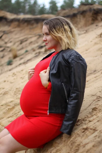 Mladá těhotná dívka v červených šatech na písku. těhotenství je radost. Velké těhotné břicho. nový život. — Stock fotografie