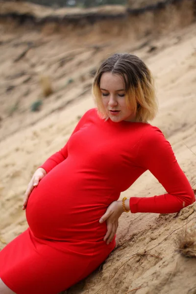 Junge schwangere Mädchen in einem roten Kleid auf dem Sand. Schwangerschaft ist eine Freude. dicker Schwangerschaftsbauch. Neues Leben. — Stockfoto