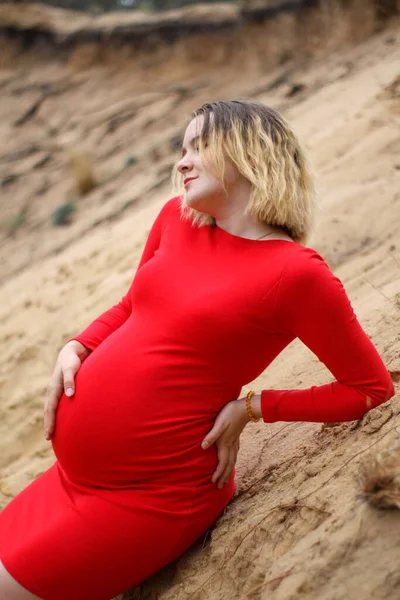 Jovem menina grávida em um vestido vermelho na areia. gravidez é uma alegria. barriga grande grávida. nova vida. — Fotografia de Stock