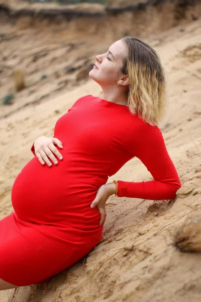 Jovem menina grávida em um vestido vermelho na areia. gravidez é uma alegria. barriga grande grávida. nova vida. — Fotografia de Stock