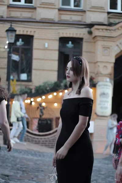 Dziewczyna w czarnej krótkiej sukience na wieczornej ulicy. na tle światła, sklepy. — Zdjęcie stockowe