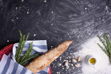 French baguette with ingredients: rosemary, olive oil, garlic and flour over black grunge background. Top view, copy space.