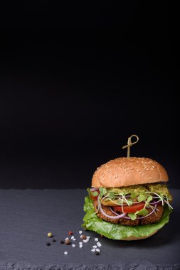 Juicy homemade hamburger with grilled beef cutlet, pepper, salad, tomato, mushrooms, onion surved with pepper and sea salt on black stone board, space for text.