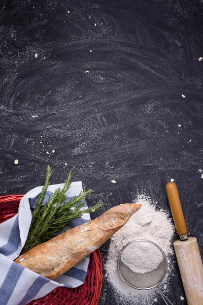 Baguette integrale in cesto rosso con rosmarino, farina e mattarello su fondo grunge nero. Vista dall'alto, spazio di copia, verticale . — Foto Stock