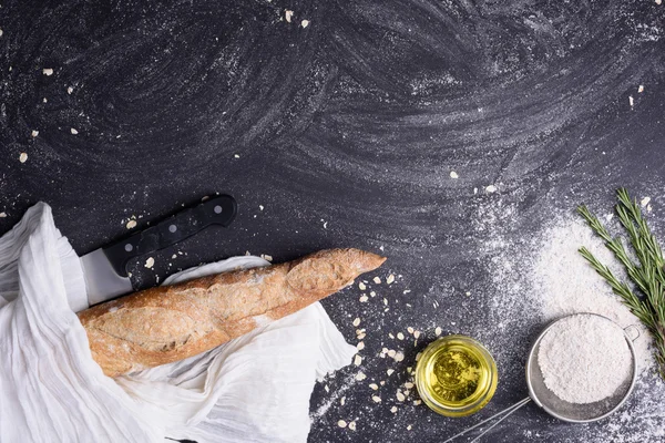French baguette with ingredients: rosemary, olive oil, garlic, knife and flour over black grunge background. Top view, free text space. — Stock Photo, Image