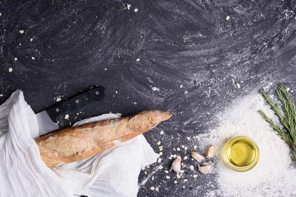 Baguette fresca con rosmarino, olio d'oliva, aglio, coltello e farina su fondo grunge nero. Vista dall'alto, copia spazio . — Foto Stock