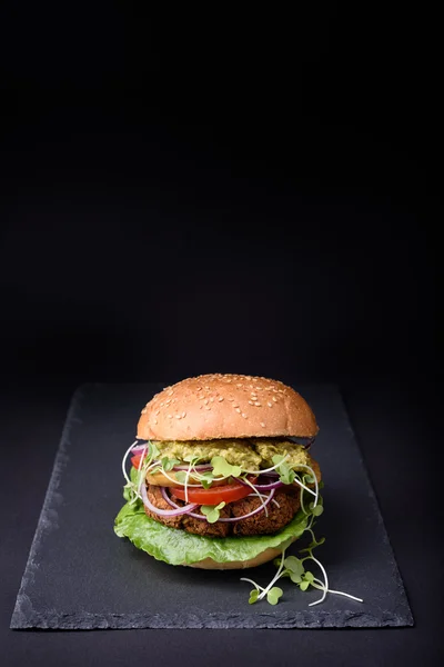 Veggie falafel burger with pesto sauce, tomato, cress salad and onion on black slate board. Space for text.