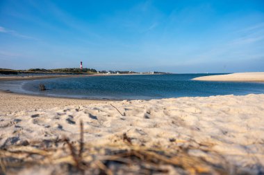 Deniz fenerli sahil. Uzakta deniz feneri olan bir körfezin etrafında kumlu bir sahil kıvrılır. Sakin sular berrak gökyüzünü yansıtır.