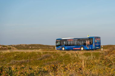 Almanya 2 Ekim 2025. Intersport reklamı olan mavi bir şehir otobüsü açık bir gökyüzünün altında kum tepelerinin içinden geçiyor. Otobüs, doğal kıyı manzarasıyla çelişiyor..
