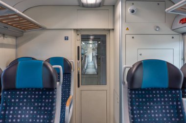 Uzun bir tren koridoru modern vagonlar boyunca uzanır ve mavi koltuklar desenlidir. İlerideki kapı bir sonraki bölüme açılıyor..