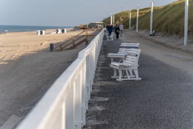Banklarla dolaş. Beyaz banklar kumlu sahile bakan sahil şeridini kaplıyor. İnsanlar arka planda berrak bir gökyüzünün altında yürür.