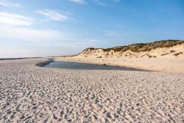 Dar bir gelgit havuzu kumlu sahil boyunca ilerler, yukarıdaki mavi gökyüzünü yansıtır. Çevreleyen kum tepeleri yavaşça arka planda yükselir.