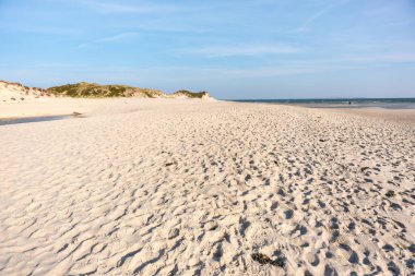 Uçsuz bucaksız kumlu sahil mavi gökyüzünün altında denize açılıyor. Uzakta hafif dalgalar görülebiliyor. Huzurlu manzara rahatlamaya davetiye çıkarır..