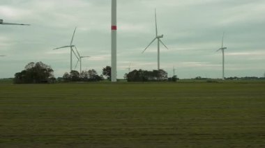 Rüzgar türbinleri bulutlu gökyüzünün altında engin yeşil bir alanda yavaşça döner. Video, hareket halindeki bir trenden görülen düz bir kırsal alanda yenilenebilir enerji yapılarını gösteriyor..