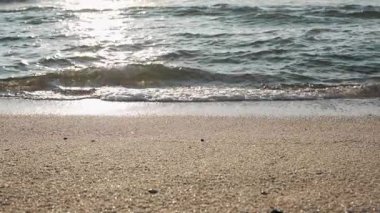 Küçük dalgalar kıyıya vururken yumuşak güneş ışığı kumda parlıyor. Yavaş çekim ışık ve suyun oyununu yakalar..