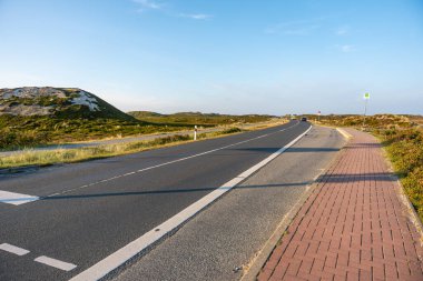 Coastal Road and Bus Stop. A quiet coastal road curves through the dunes, lined with a red-brick sidewalk. The golden hour light casts long shadows and warmth.