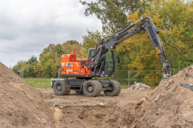 Almanya 11 Ekim 2025. Parlak turuncu bir kazıcı toprak yığınlarıyla çevrili bir inşaat sahasında duruyor. Makineler hidrolik kol ve büyük lastikler yeşil sonbahar ağaçlarıyla tezat oluşturuyor..
