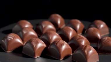 Çikolata aranjmanı. Sıralar dolusu parlak çikolatalı pralinler siyah bir yüzey üzerinde pürüzsüz bir şekilde döner. Her parça ışığı güzel yansıtıyor, lüksü ve hassasiyeti vurguluyor..