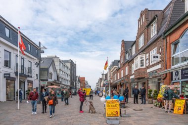 Almanya Sylt 2 Ekim 2025. İnsanlar bulutlu bir gökyüzünün altında butikler ve kafelerle dolu canlı bir yaya caddesinde yürüyor. Bayraklar ve mağaza tabelaları kasabanın atmosferini güzelleştirir..