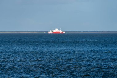 Almanya Sylt 27 Ekim 2025. Bir kırmızı ve beyaz yolcu feribotu uzak bir ada kıyısı yakınlarındaki sakin mavi sularda yol alır. Gemi deniz ve gökyüzü ile keskin bir tezat içinde..