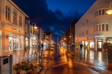 Almanya Sylt 27 Ekim 2025. Alacakaranlıkta ışıldayan mağazalar ve ıslak yansımalarla dolu canlı bir alışveriş caddesi. İnsanlar altın sokak lambaları altında yürür.