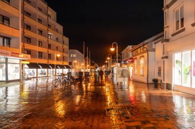 Almanya Sylt 28 Ekim 2025. Geceleri sıcak sokak lambaları altında yağmurdan sonra bir şehir caddesi parlar. Islak kaldırım taşlarında yansımalar parıldıyor insanlar aydınlanmış dükkanlarda yürürken..