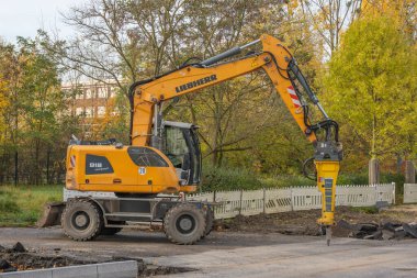 Almanya 31 Ekim 2025. Liebherr kompakt tekerlekli kazıcı hidrolik şalter ile yol yüzeyinde çalışıyor. Makine sonbahar ağaçlarının ve beyaz engellerin önünde duruyor..