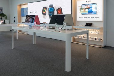 Almanya 31 Ekim 2025. Modern bir Apple mağazası, gösterişli beyaz masalarda iMac ve aksesuarlar sergiliyor. Kurulum temiz tasarım ve premium teknolojisini vurguluyor.