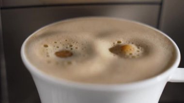 Bir fincan taze kahvenin üzerinde oluşan krema yakın plan görüntüsü. Yüzeyde küçük kabarcıklar dans ediyor. Ağır çekimde damlayan kahve damlaları köpüklü bir fincan espressoya dönüşür..