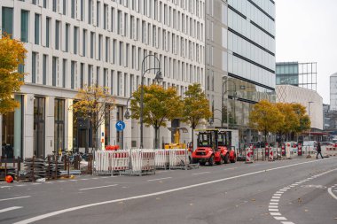 Bariyerler ve kırmızı forkliftlerle inşa edilmiş sokak bölümü. Modern faşatlar ve hizalı ağaçlar eğriyi çiziyor.