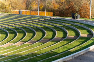 Eğri büğrü amfitiyatro merdivenleri tekrar eden çimen ve beton hatlarını süpürüyor. Uzun gölgeler minimalist sahneye ritim katar.