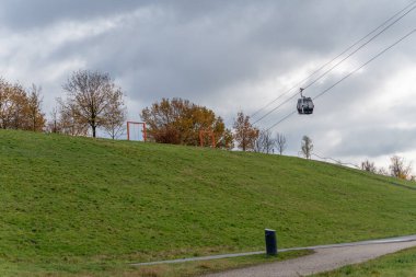 Bir tramvay şehir parkında çimenli bir yamacın üzerinde süzülüyor. Sonbahar ağaçları ve turuncu çerçeveler dramatik bulutlu bir gökyüzüne karşı duruyor.