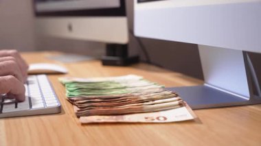 Birisi yazarken, uzun avroluk banknotlar ahşap bir masanın üzerine bilgisayar monitörlerinin önünde yayılıyor. Kamera para boyunca sağdan sola hareket eder..