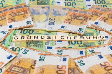 Grundsicherung ahşap kiremitlerin içinde karışık avro banknotları arasında görünüyor. Refah desteği, yaşam maliyeti ve kamu yardımı hakkındaki sembolik yaşam.