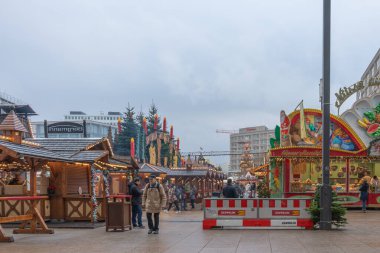 Almanya 3 Aralık 2025. Berlin 'de kış manzaralı ahşap tezgahlar, ışıklar ve ziyaretçiler. Bulutlu gökyüzü festival şehir meydanı ve tatil atmosferini çerçeveletiyor.