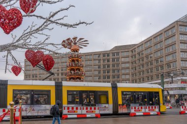 Almanya 3 Aralık 2025. Sarı tramvay, kırmızı kalp ışıklarıyla süslenmiş bir Berlin Noel pazarından geçiyor. Kış sokak sahneleri toplu taşıma, şenlik tezgahları ve yayaları birleştiriyor.
