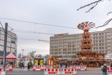 Almanya 3 Aralık 2025. Büyük ahşap bir Noel piramidi ve çizgili çadırları olan Berlin Noel pazarının manzarası. Kış meydanında inşaat bariyerleri ve tramvay kabloları var..