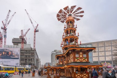 Almanya 3 Aralık 2025. Berlin 'de bir şehir pazarında Noel piramidi. Arkasında vinç ve inşaat var. Sarı bir tramvay ve yayalar şehrin kış koşuşturmasını gösteriyor..