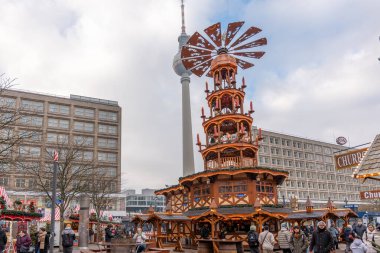 Almanya 3 Aralık 2025. Berlin Noel piyasa sahnesi. Arkasında büyük bir ahşap piramit ve televizyon kulesi var. Kış meydanını kabinler, ışıklar ve ziyaretçiler doldurdu.