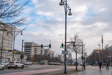 Almanya 3 Aralık 2025. Berlin kavşağında trafik ışıkları, arabalar ve yayalar bulutlu bir kış gökyüzü altında. Şehir altyapısını modern sokak lambaları ve bisiklet yolları vurguluyor.
