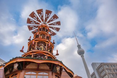 Berlin TV Kulesi 'nin arka planında parıldayan bir Noel piramidinin alçak açılı görüntüsü. Mavi gökyüzü ve bulutlar dramatik bir kış şehri havası ekler..