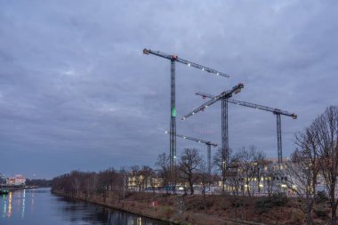 İnşaat vinçleri nehir kıyısındaki bir şantiyenin üzerinde alacakaranlık gökyüzünün altında yükseliyor. Kanal ve çıplak ağaçlar sert bir şehir kışı manzarası yaratır..