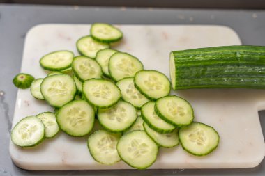 Taze salatalık dilimleri beyaz bir kesme tahtasının üzerinde kısmen kesilmiş bir salatalığın yanında yatar. Parlak yeşil kabuk ve sulu merkezler taze ve sağlıklı malzemeler olduğunu gösteriyor..