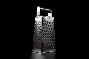 Siyah arka planda duran paslanmaz çelik rendesi ve dramatik stüdyo ışıklandırması. Temiz, minimalist mutfak aleti görüntüsü dokuyu ve yansımaları vurguluyor.