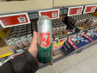Almanya 29 Aralık 2025. Süpermarket içecek reyonunda uzun bir bira kutusunu tutarken. Fiyat etiketleri ve istiflenmiş kutular perakende alışveriş ve tüketici seçimini vurguluyor.